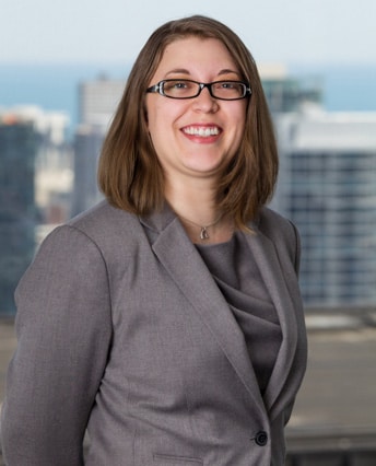 Female attorney in gray blazer smiling, wearing glasses, with Chicago skyline and lake in background