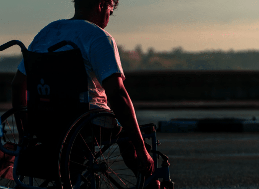 Person in a wheelchair facing a sunset horizon, viewed from behind in dramatic low lighting