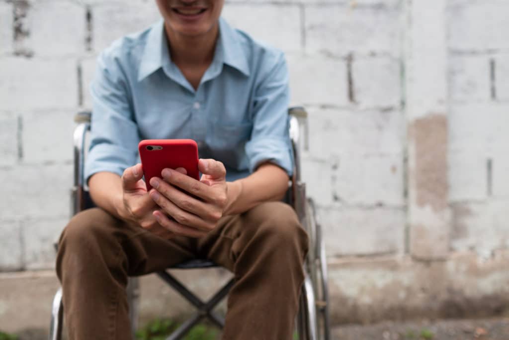Smiling person in a wheelchair using a red smartphone outdoors near a brick wall