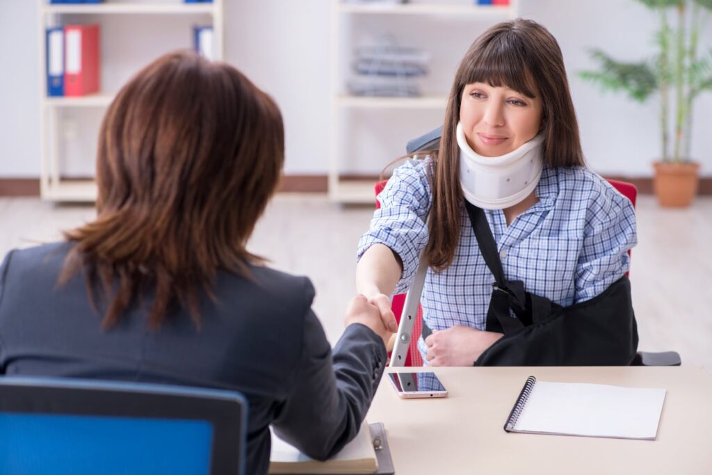 Injured woman wearing a neck brace and arm sling shaking hands with a personal injury attorney during a legal consultation