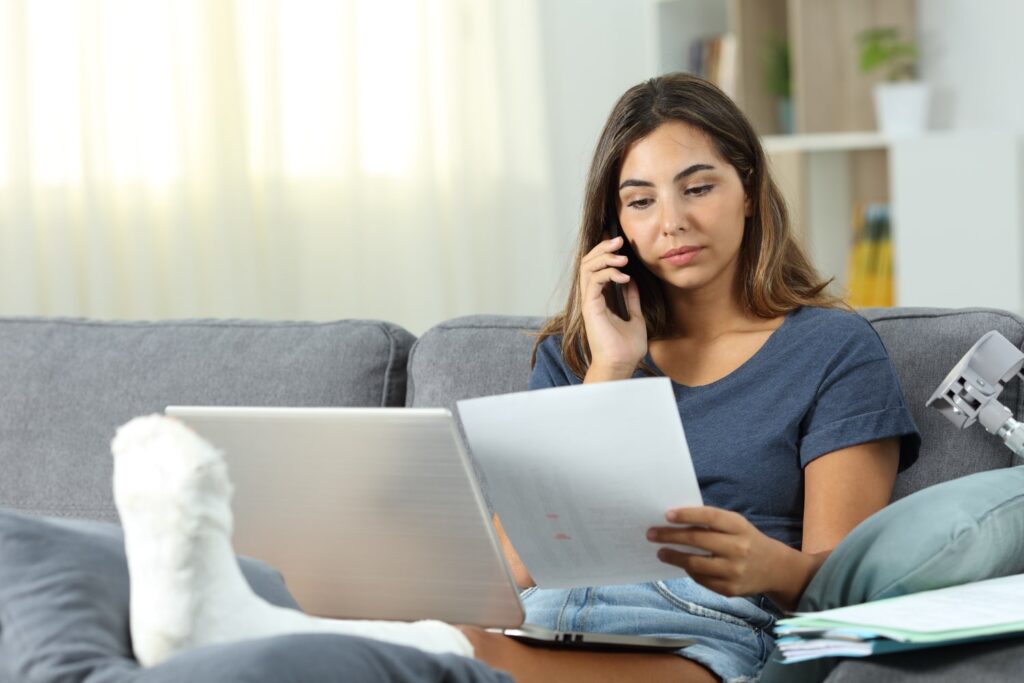 Woman with a broken foot in a cast reviewing legal documents and talking on the phone while seated on a couch at home