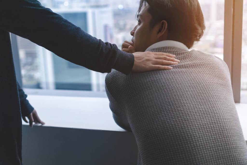 A person placing a comforting hand on a distressed man's shoulder as he gazes out a window