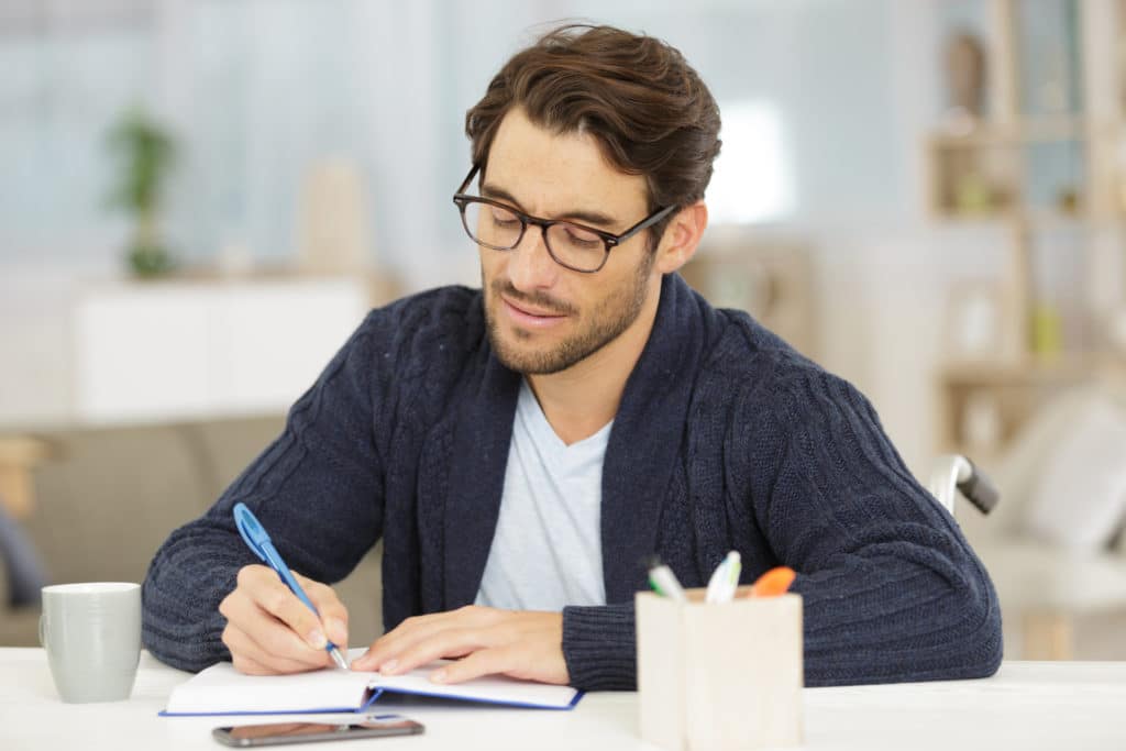 Young man with glasses writing notes in a notebook at a desk with a coffee mug and pen holder nearby