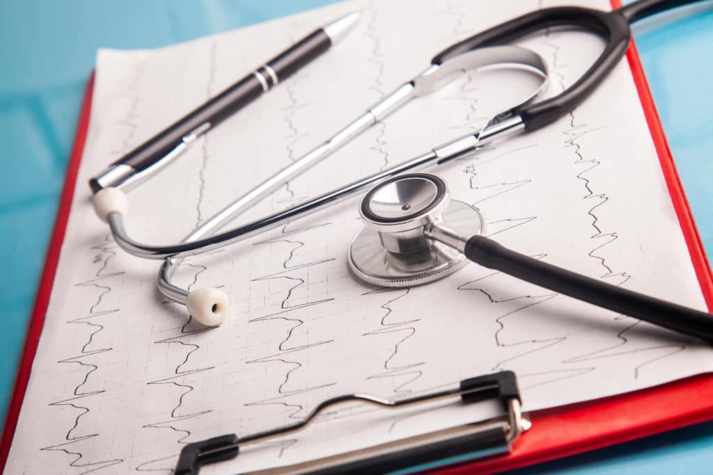 Stethoscope and pen resting on EKG heart rhythm printout attached to a red medical clipboard