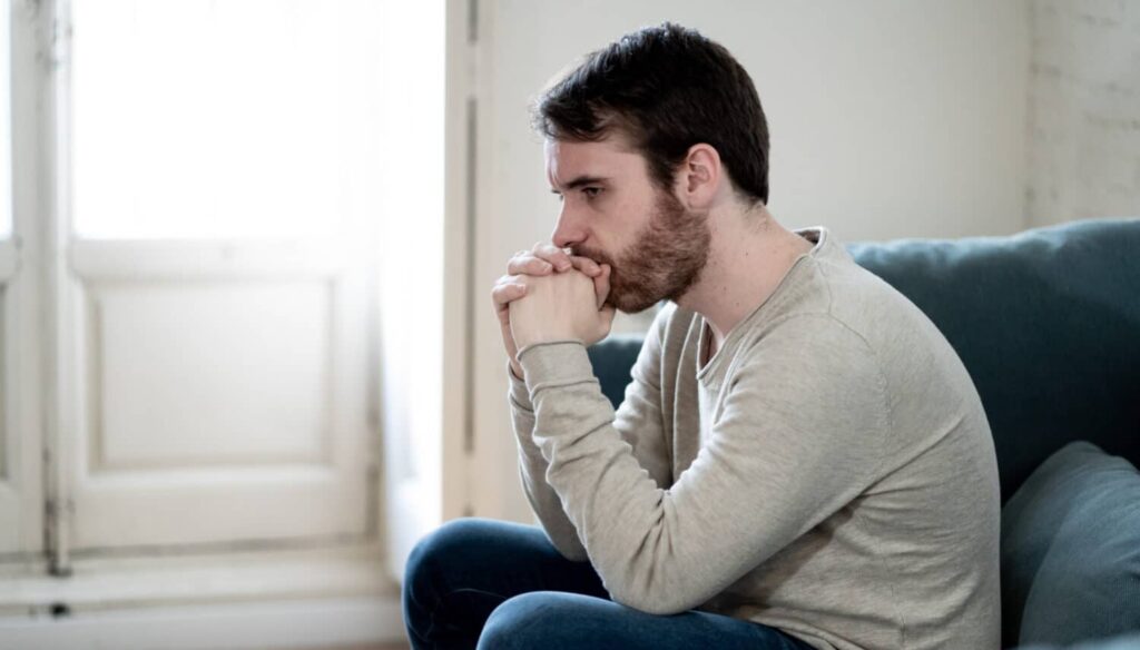 Young man sitting on a couch with hands clasped, looking distressed and deep in thought near a bright window