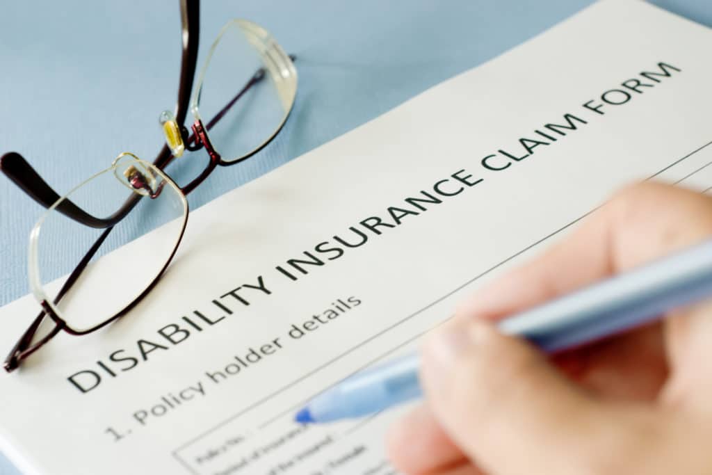 Person completing a disability insurance claim form with a pen, glasses resting nearby on the document