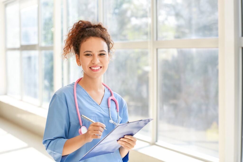 Smiling female nurse in blue scrubs with pink stethoscope holding clipboard and pen in a bright medical facility hallway