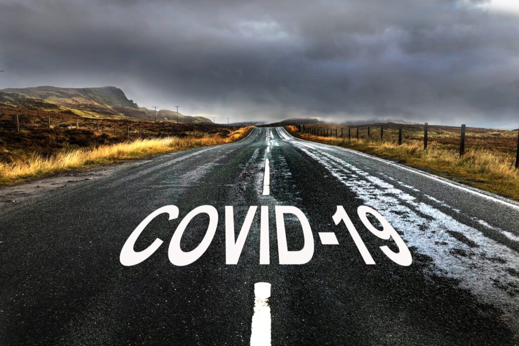 Empty rural road under stormy skies with "COVID-19" painted on the pavement, symbolizing pandemic challenges ahead
