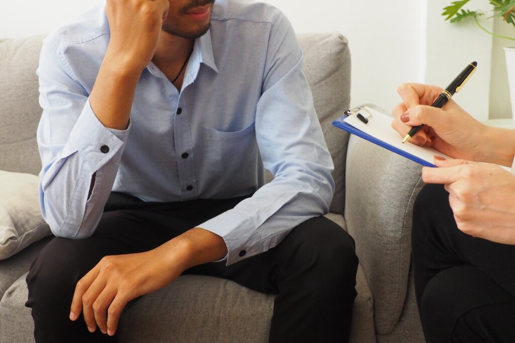 Distressed man sitting on a couch during a consultation while a professional takes notes on a clipboard