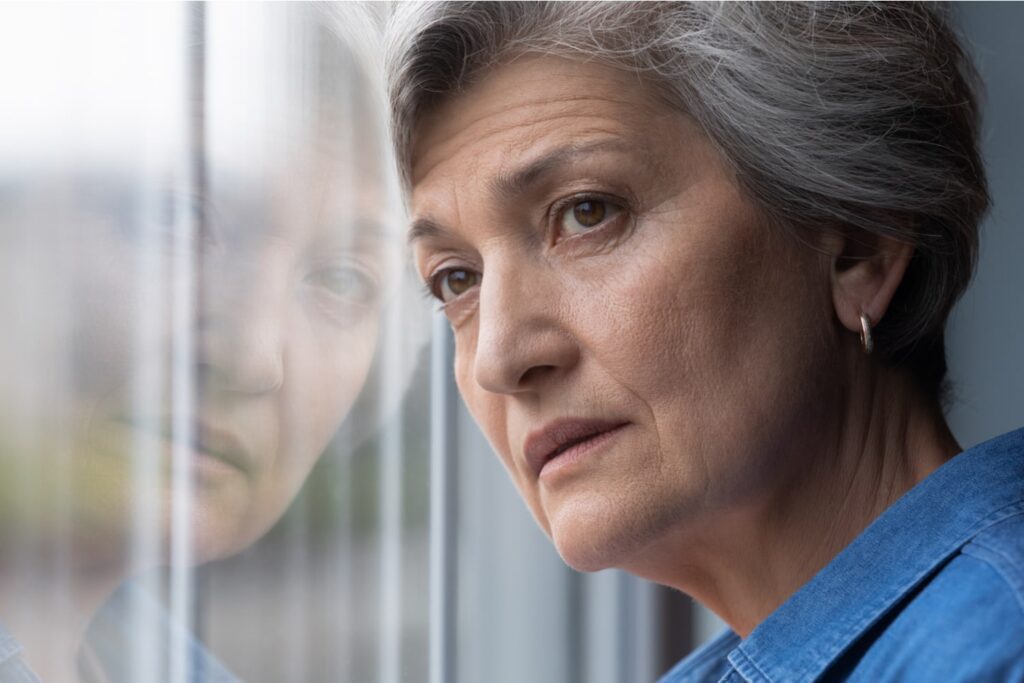 Older woman with gray hair gazing pensively out a window, her reflection visible in the glass