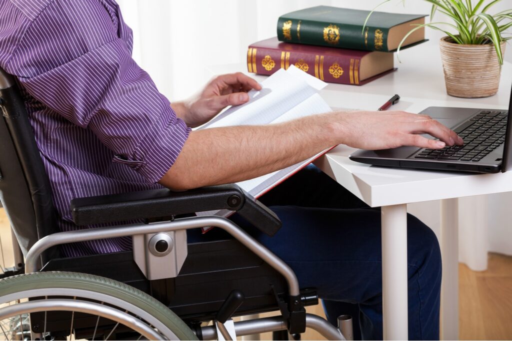Man in wheelchair reviewing documents and using laptop at desk with stacked law books, researching disability benefits