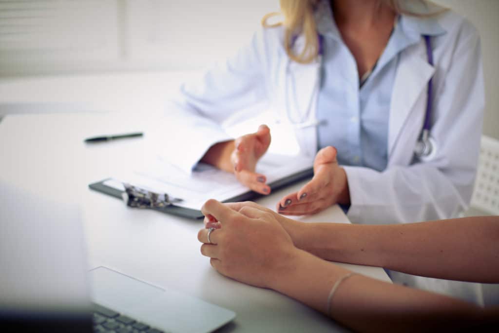 Doctor in white coat with stethoscope gesturing while consulting a patient across a desk with clipboard and documents