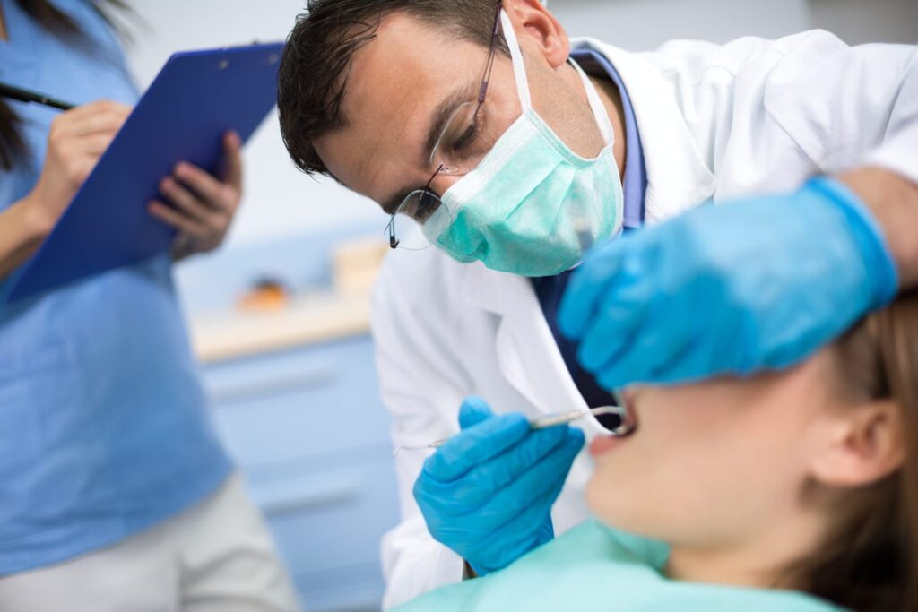 Dentist in mask and gloves performing a dental procedure on a patient while an assistant records notes on a clipboard