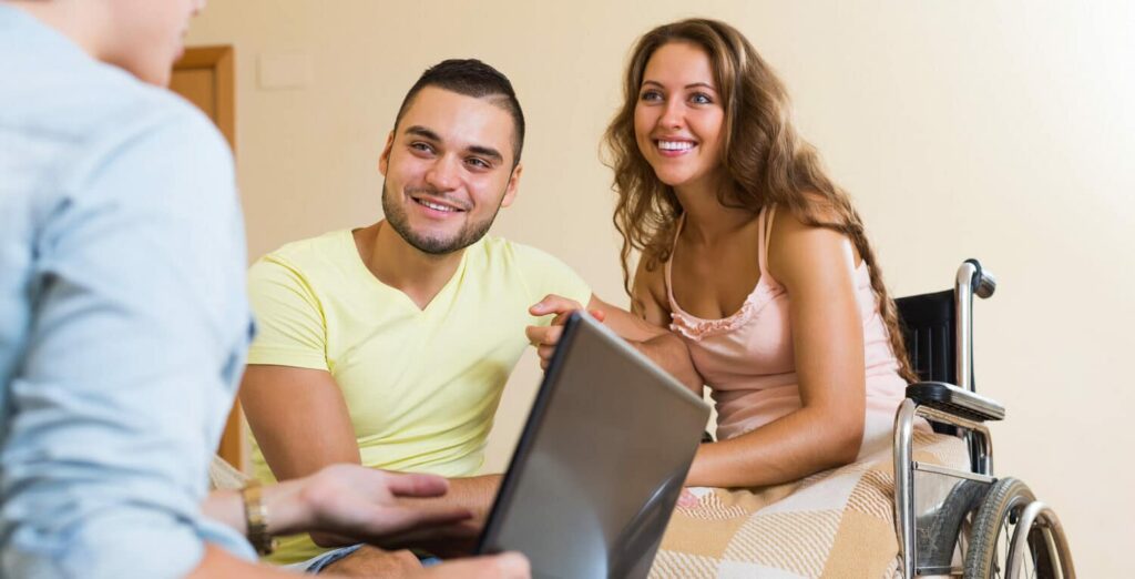 A smiling couple meets with an advisor holding a laptop; the woman is seated in a wheelchair during the consultation