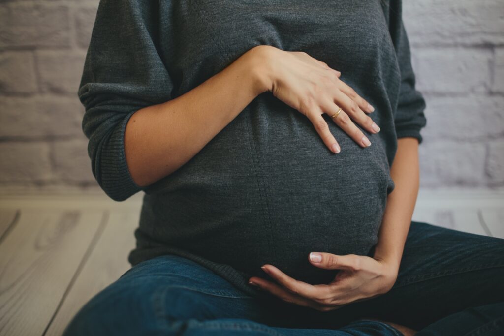Pregnant woman in gray sweater and jeans gently cradling her baby bump with both hands while seated indoors