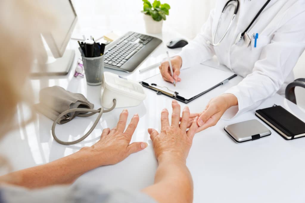 Doctor examining an elderly patient's hands during a medical consultation while taking notes on a clipboard