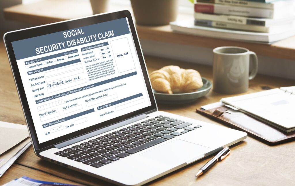 Laptop displaying a Social Security Disability Claim form on screen, placed on a wooden desk with a notebook and pen nearby