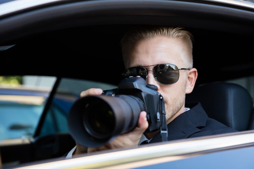 Private investigator in sunglasses conducting covert surveillance from a vehicle using a professional telephoto camera lens