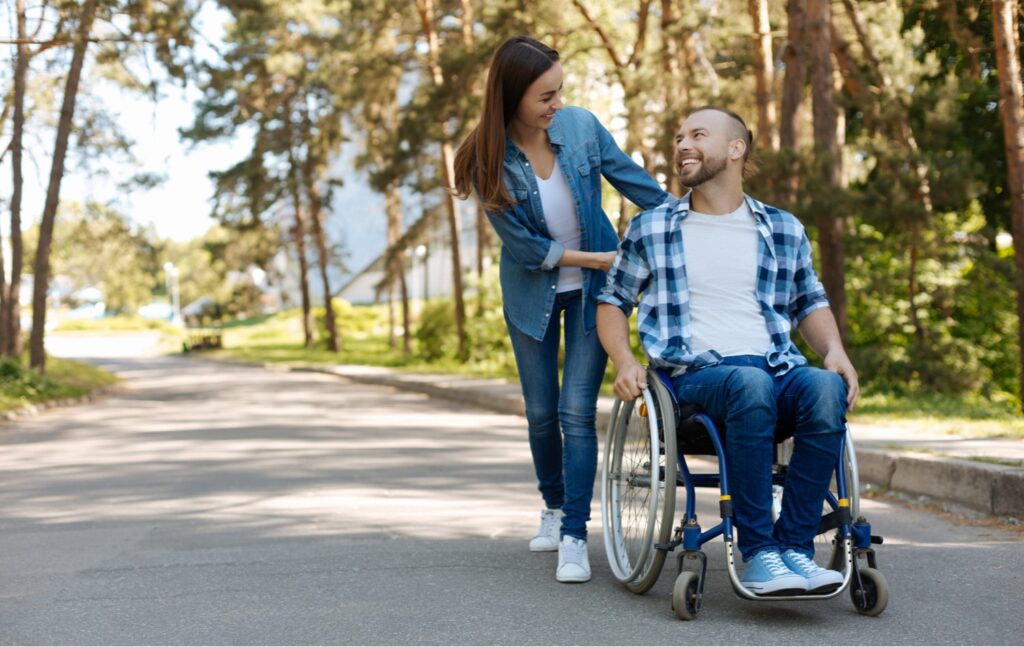 Woman helping a smiling man in a wheelchair along a sunny tree-lined park path, representing disability support and care