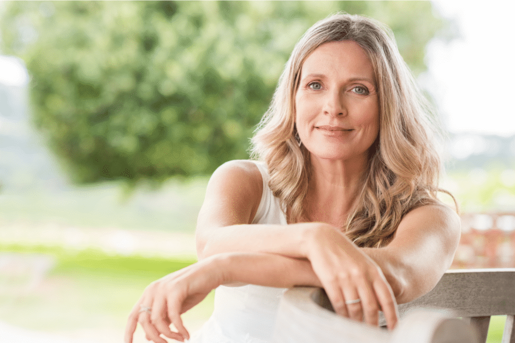 Middle-aged woman with blonde hair sitting relaxed on an outdoor bench, smiling calmly against a green background