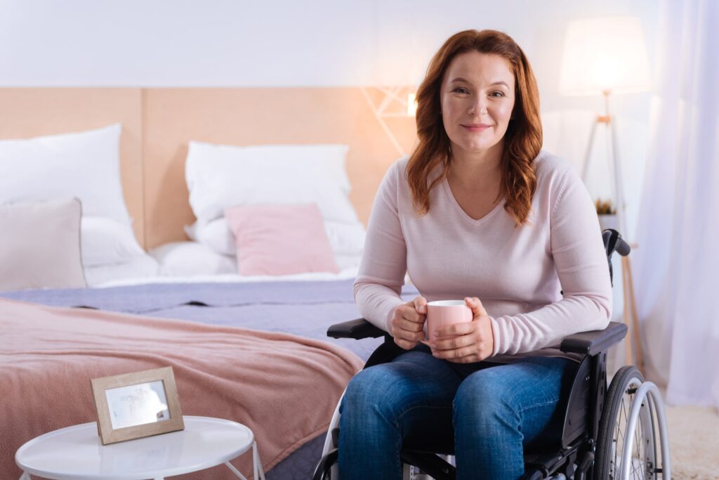 Woman in a wheelchair holding a coffee mug, smiling calmly in a cozy bedroom setting