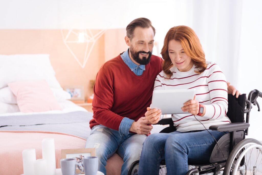 Man and woman in a wheelchair smiling while reviewing information on a tablet together in a home setting