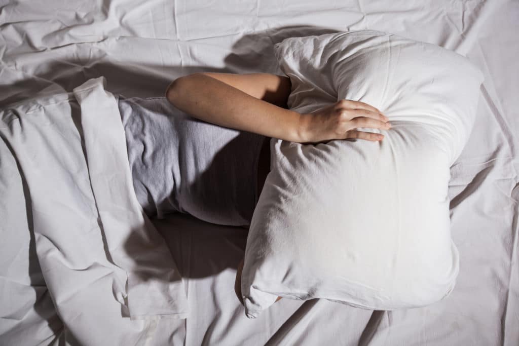 Person lying in bed with face hidden under a pillow, depicting sleep disturbance or emotional distress